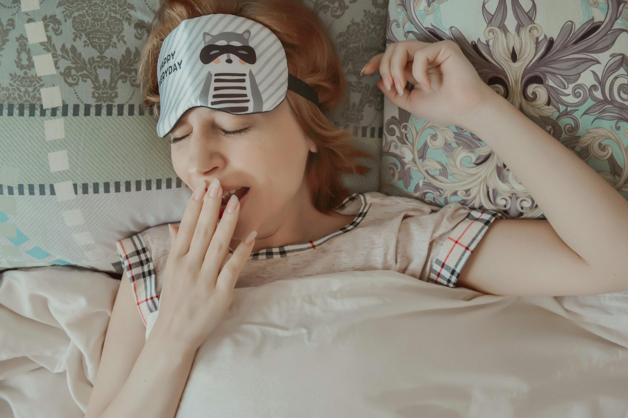 A woman in cozy pajamas yawning while waking up in bed, wearing a sleep mask.