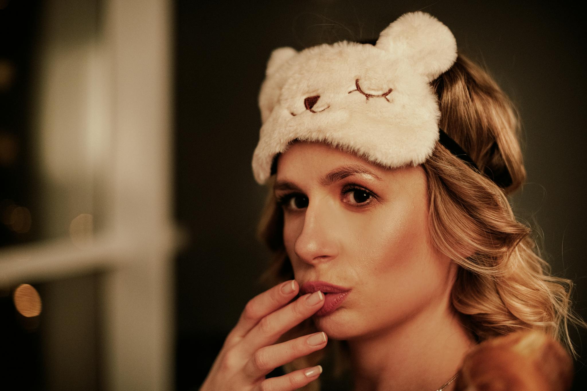 A woman wearing a cute bear-themed sleep mask poses thoughtfully indoors.